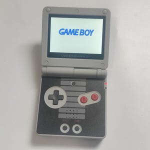 May include: A gray Game Boy Advance SP handheld game console, open to display the words "GAME BOY" in blue on the screen. The console features a directional pad, red A and B buttons, and a black and gray design.