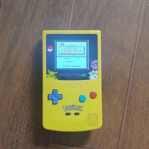 May include: A yellow Game Boy Color handheld video game console with a Pikachu design. The screen shows the game "Super Mario Land" with the text "GAME BOY COLOR" on the bottom of the screen.