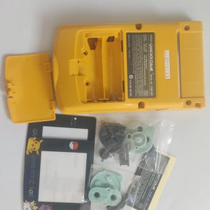 K&ouml;nnte beinhalten: Eine gelbe Game Boy Color Handheld-Spielkonsole, mit ge&ouml;ffnetem Batteriefach. Die Konsole wird von einer schwarz-wei&szlig;en Karte mit Pikachu-Grafiken und verschiedenen Ersatzteilen begleitet.