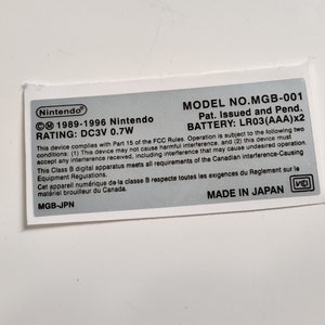 May include: A white label with black text and a silver background. The label has the text "Nintendo 1989-1996 Nintendo RATING: DC3V 0.7W MODEL NO.MGB-001 Pat. Issued and Pend. BATTERY: LR03 (AAA)x2 This device complies with Part 15 of the FCC Rules. Operation is subject to the following two conditions: (1) This device may not cause harmful interference, and (2) this device must accept any interference received, including interference that may cause undesired operation. This Class B digital apparatus meets all requirements of the Canadian interference-Causing Equipment Regulations. Cet apparell numérique de la classe B respecte toutes les exigences du Reglement sur le matériel brouilleur du Canada. MGB-JPN MADE IN JAPAN"