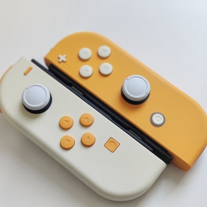 Puede incluir: Un mando de Nintendo Switch blanco y amarillo con botones blancos y una carcasa amarilla. El mando est&aacute; dividido en dos, con el lado izquierdo blanco y el lado derecho amarillo.