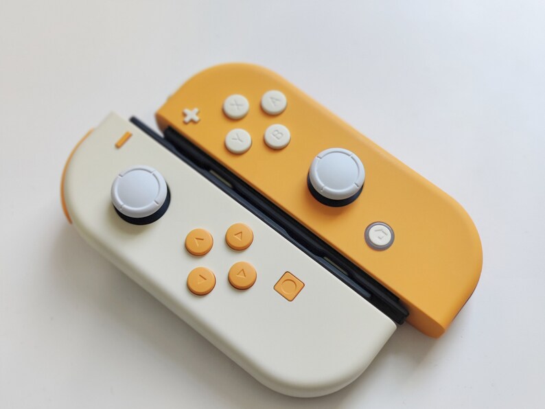 Puede incluir: Un mando de Nintendo Switch blanco y amarillo con botones blancos y una carcasa amarilla. El mando est&aacute; dividido en dos, con el lado izquierdo blanco y el lado derecho amarillo.