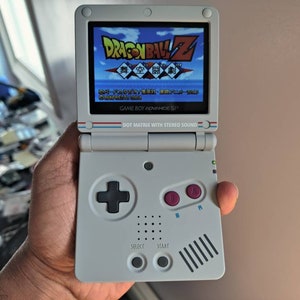 May include: A white Game Boy Advance SP handheld gaming console with a black and red button and a screen displaying the Dragon Ball Z video game.