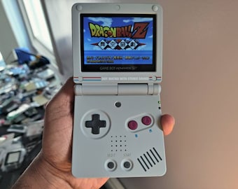 Puede incluir: Una consola de juegos portátil Game Boy Advance SP blanca con un botón negro y rojo y una pantalla que muestra el videojuego Dragon Ball Z.