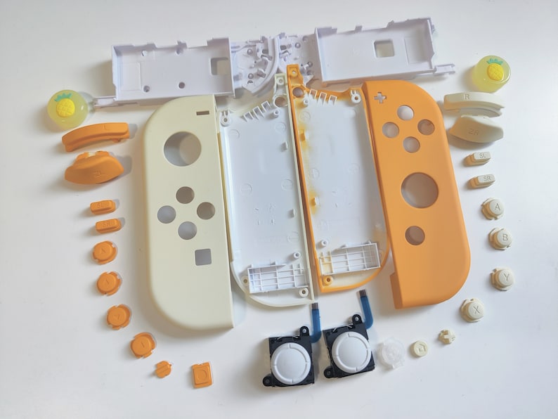 Puede incluir: Un conjunto de piezas de repuesto para una consola Nintendo Switch, que incluye una carcasa blanca y naranja, botones y joysticks. Las piezas est&aacute;n dispuestas sobre una superficie blanca.