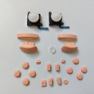 Puede incluir: Un conjunto de botones de repuesto para un mando de videojuegos. El conjunto incluye una variedad de botones de diferentes formas y tamaños, incluidos botones direccionales, botones de cara y botones de hombro. Los botones son todos de color melocotón claro.