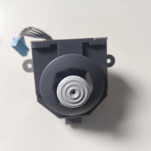 Puede incluir: Un componente electrónico gris oscuro con una perilla blanca estriada circular en el centro. El componente tiene una base hexagonal y un conector azul con cables. Se ven dos pequeñas pestañas metálicas en los lados.