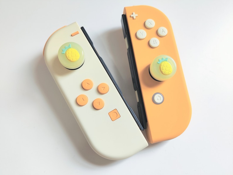 Puede incluir: Dos mandos Joy-Con para Nintendo Switch con tapas de botones en forma de pi&ntilde;a. Un mando es blanco con botones naranjas y el otro es naranja con botones blancos.