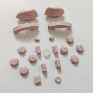 May include: A set of pink replacement buttons for a gaming console. The set includes buttons labelled 'ZL', 'ZR', 'L', 'R', 'A', 'B', 'X', 'Y', 'SL', 'SR', and directional arrows.