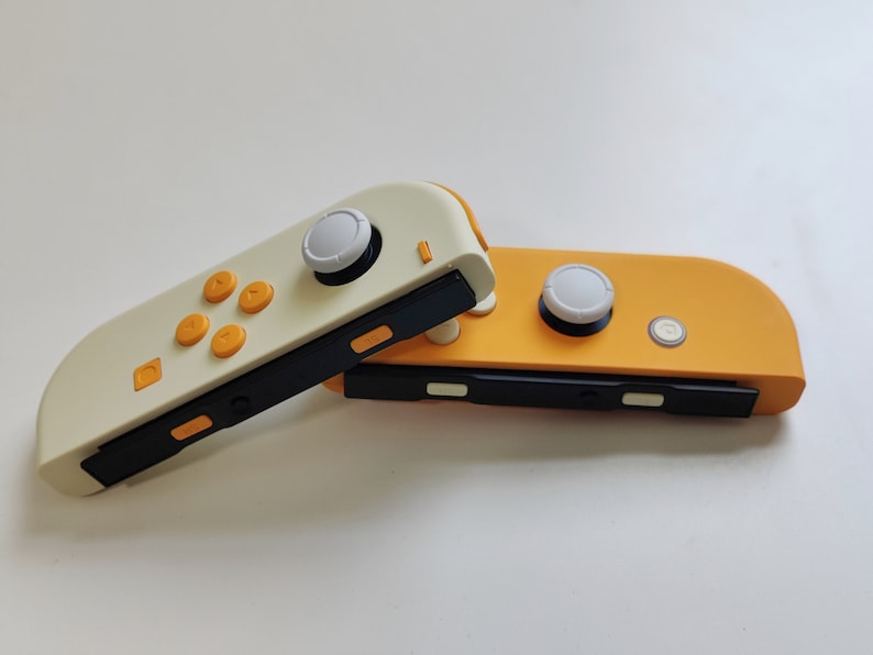 Puede incluir: Dos mandos Joy-Con para Nintendo Switch, uno de color crema con botones naranjas y el otro de color naranja brillante con botones blancos.