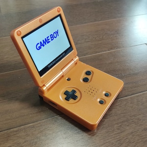 May include: An orange Game Boy Advance SP handheld gaming console with a blue "GAME BOY" logo on the screen. The console has a black directional pad and buttons.