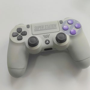 May include: A grey PlayStation 4 controller with purple buttons. The controller has the text "SUPER STATION" printed on the front.