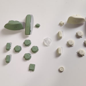 May include: A set of replacement buttons for a video game controller. The buttons are in two colors: green and white. The green buttons are labeled "ZL", "ZR", "SL", "SR", "L", and "R". The white buttons are labeled "A", "B", "X", "Y", and "Start".