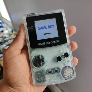 Può includere: Una console per videogiochi portatile Game Boy Color verde traslucida con uno schermo nero che mostra il testo "GAME BOY".