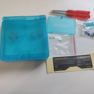 May include: A blue Game Boy Advance SP handheld gaming console with a clear plastic shell. The console is open and the screen is visible. The console is surrounded by various parts and accessories, including a clear plastic faceplate, a small bag of white plastic buttons, a small bag of screws, and a sticker with the text "GAME BOY ADVANCE SP" and "KJH111318460".