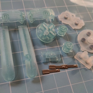 May include: A set of translucent green replacement buttons for a video game controller. The set includes a directional pad, four face buttons, two shoulder buttons, two trigger buttons, and two small metal pieces.