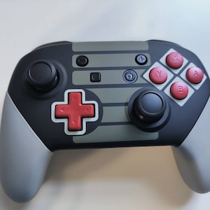 May include: A black and gray video game controller with red buttons. The controller has a gray and white striped design on the top and a black and gray design on the bottom. The controller has a plus sign button, a home button, and a joystick.