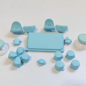 Puede incluir: Un conjunto de botones de repuesto de color azul claro para un mando de videojuegos. El conjunto incluye dos sticks analógicos, cuatro botones frontales, dos botones de hombro, dos gatillos y una almohadilla direccional.