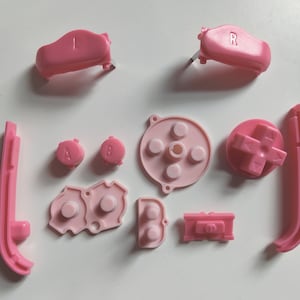 Puede incluir: Un conjunto de botones de repuesto de color rosa para una consola de juegos. El conjunto incluye botones etiquetados como 'A', 'B', 'L', 'R' y 'Start', así como un pad direccional y un pad de control de cuatro direcciones.