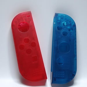 May include: A pair of transparent red and blue game controller shells. The red shell is on the left, and the blue shell is on the right. Both shells have visible internal components and button placements, with a glossy finish.