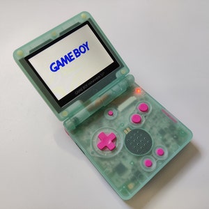 Puede incluir: Una consola de juegos portátil Game Boy Advance SP verde menta, abierta para mostrar la palabra "GAME BOY" en azul en la pantalla. La consola tiene botones rosas y una cruceta rosa.