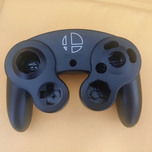 May include: A black GameCube controller with a white Super Smash Bros. logo on the top. The controller has a black and white D-pad, two black buttons, and two black analog sticks.