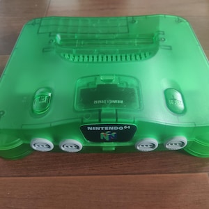 Könnte beinhalten: Eine transparente, grüne Nintendo 64-Spielkonsole. Die Konsole hat eine rechteckige Form mit abgerundeten Kanten und das Nintendo 64-Logo. Sie hat vier Controller-Anschlüsse und mehrere Tasten.