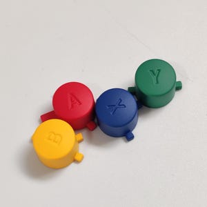 Op de afbeelding: Vier ronde plastic knoppen met letters en symbolen. De knoppen zijn rood, geel, blauw en groen. De letters op de knoppen zijn A, B, X en Y.