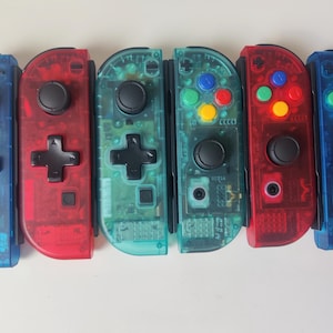 Może przedstawiać: Sześć kolorowych kontrolerów Nintendo Switch Joy-Con. Kontrolery są przezroczyste i mają przyciski w różnych kolorach. Kontrolery są ułożone w rzędzie od lewej do prawej: niebieski, czerwony, zielony, zielony, czerwony, niebieski.