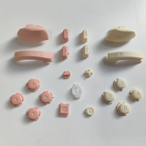 Op de afbeelding: Een set vervangende knoppen voor een gameconsole. De knoppen zijn in twee kleuren: crème en lichtroze. De knoppen zijn gelabeld met letters en symbolen, waaronder "A", "B", "X", "Y", "L", "R", "Start", "Select" en pijltjes.