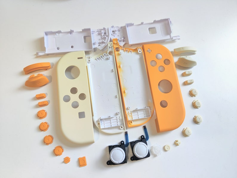 Puede incluir: Un conjunto de piezas de repuesto para un mando de Nintendo Switch. Las piezas son de pl&aacute;stico blanco y naranja e incluyen los botones frontales, los botones de los hombros, los joysticks y una placa posterior.
