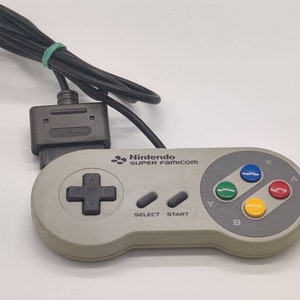 Könnte beinhalten: Ein grauer Super Nintendo Entertainment System-Controller mit einem schwarzen Kabel. Der Controller hat einen grau-schwarzen Steuerkreuz, vier farbige Knöpfe und den Text "Nintendo SUPER FAMICOM" auf der Oberseite.