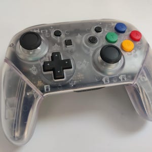 May include: A clear, translucent Nintendo Switch controller with black buttons and joysticks. The face buttons are coloured red, green, yellow, and blue.