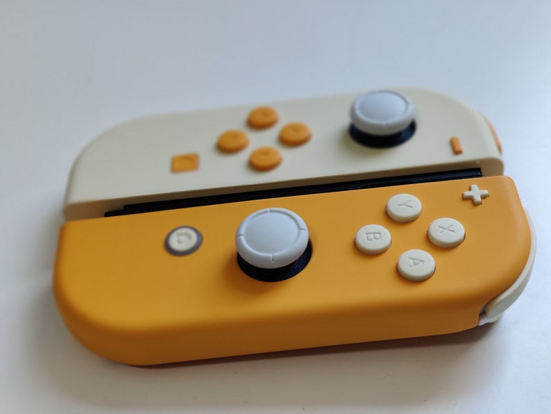 Puede incluir: Un mando de Nintendo Switch amarillo y blanco con botones blancos y una palanca de mando blanca. El mando tiene la parte superior amarilla y la inferior blanca.