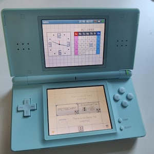 May include: A light blue handheld gaming console with dual screens. The top screen displays a clock and a calendar for February 2000. The bottom screen shows the main menu with icons for PictoChat and DS Download Play, along with system status text.