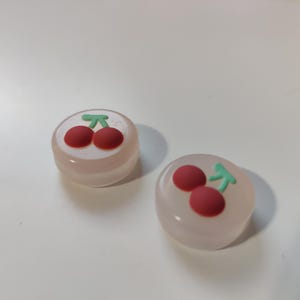 Puede incluir: Dos pequeños tapones de teclas redondos de silicona rosa translúcida con un diseño de cereza. Cada tapón de tecla presenta dos cerezas rojas con tallos y hojas verdes.