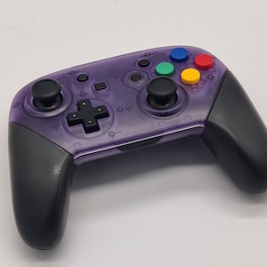May include: A purple translucent Nintendo Switch controller with black buttons and joysticks. The controller has colored buttons on the top.