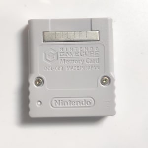 Könnte beinhalten: Eine hellgraue Nintendo GameCube Speicherkarte. Auf der Karte sind das Nintendo GameCube-Logo, die Worte "Memory Card" und "MADE IN JAPAN" aufgedruckt. Außerdem das Nintendo-Logo.