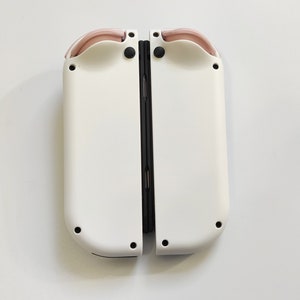 Puede incluir: Un mando de juego Nintendo Switch Joy-Con blanco con detalles en rosa. El mando est&aacute; cerrado y las dos mitades est&aacute;n conectadas por una bisagra negra.