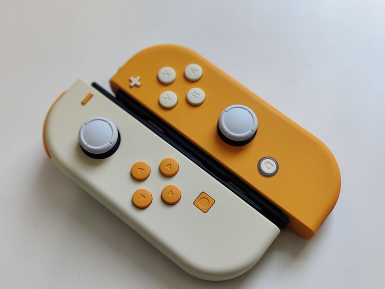 Puede incluir: Un mando de juego Nintendo Switch Joy-Con blanco y amarillo con botones blancos y un bot&oacute;n de inicio blanco. El mando est&aacute; en una configuraci&oacute;n dividida, con el lado izquierdo que presenta una palanca anal&oacute;gica blanca y cuatro botones direccionales naranjas, y el lado derecho que presenta una palanca anal&oacute;gica blanca y cuatro botones de cara blancos.