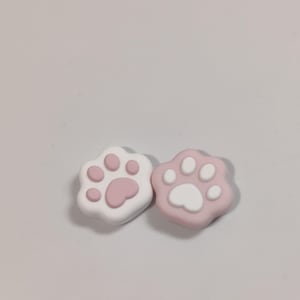 May include: Two paw-shaped accessories, one white with pink pads and the other pink with white pads. These small, decorative items are likely for a craft project or to embellish other objects. The paw prints are a cute, playful design.