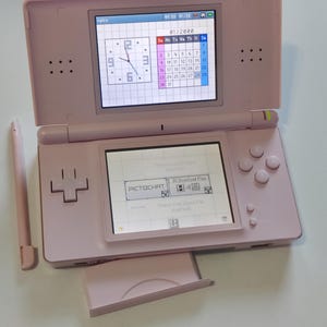 Puede incluir: Una consola de videojuegos portátil de color rosa claro con doble pantalla. La pantalla superior muestra un reloj y un calendario de enero de 2000. La pantalla inferior muestra un menú con el texto PictoChat. Un lápiz óptico rosa a juego está a la izquierda.