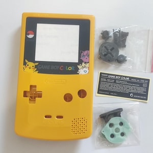 K&ouml;nnte beinhalten: Gelbe Game Boy Color Konsole mit schwarzem Bildschirm und verschiedenen Ersatzteilen. Die Konsole zeigt Pikachu und andere Pok&eacute;mon-Charaktere. Enth&auml;lt einen Aufkleber mit dem Text "GAME BOY COLOR".