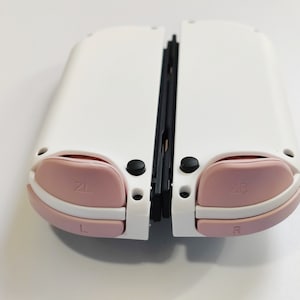 Puede incluir: Consola Nintendo Switch blanca con botones rosas y una bisagra negra. Los botones est&aacute;n etiquetados como "ZL", "ZR", "L" y "R".