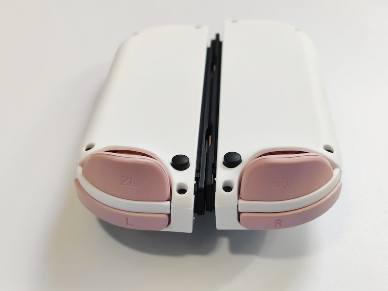 Puede incluir: Consola Nintendo Switch blanca con botones rosas y una bisagra negra. Los botones est&aacute;n etiquetados como "ZL", "ZR", "L" y "R".