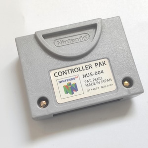 Puede incluir: Un pack de controlador gris Nintendo 64 con el texto "CONTROLLER PAK NUS-004 PAT. PEND. MADE IN JAPAN G744857 NUS-A-PA" impreso en la etiqueta.
