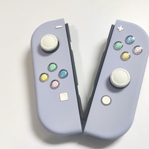 Peut inclure: Deux manettes de jeu lavande avec des joysticks blancs et des boutons multicolores. Les manettes sont ouvertes, révélant l'intérieur noir. Les boutons sont de différentes couleurs, dont le vert, le bleu, le jaune et le rose.