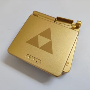 Könnte beinhalten: Ein goldener Game Boy Advance SP mit einem goldenen Triforce-Logo auf der Vorderseite. Die Konsole ist geschlossen.