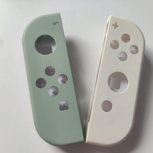 Könnte beinhalten: Zwei Controller-Schalen, eine salbeigrüne und eine cremefarbene, für eine Spielkonsole. Jede Schale hat Knopflöcher und eine kreisförmige Öffnung. Die grüne Schale hat ein Steuerkreuz und vier Knöpfe. Die cremefarbene Schale hat vier Knöpfe und einen Plus-Knopf.