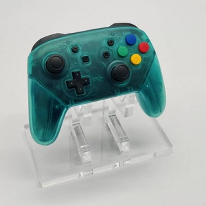 May include: A teal-colored video game controller with black buttons and a directional pad. The controller has colored buttons in red, blue, yellow, and green. It is displayed on a clear acrylic stand.